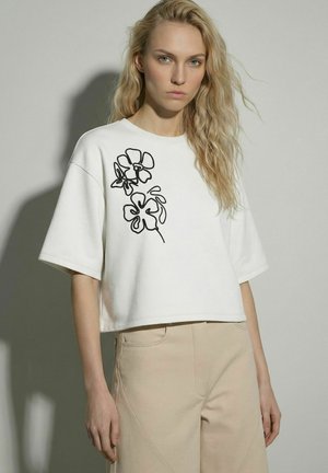 Kvinde med langt, bølget blondt hår iført hvid kort t-shirt med sort blomsterlinjekunst og beige bukser mod ensfarvet baggrund.