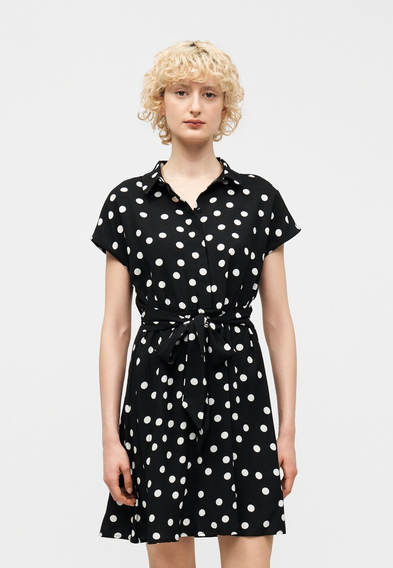 Νεαρή γυναίκα με κοντά σγουρά ξανθά μαλλιά, φορώντας μαύρο κοντομάνικο φόρεμα με λευκές πουά και φιόγκο στη μέση, που στέκεται μπροστά σε απλό φόντο.