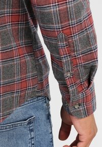 Kostkovaná flanelová košile s červenými, šedými a modrými barvami, dlouhými rukávy, knoflíkovými manžetami a zakřiveným lemem. Měkká textura s kostkovaným vzorem.