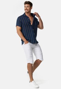 Chemise  à manches courtes bleue avec des rayures verticales blanches et dorées, associée à un short blanc jusqu'au genou et des baskets blanches. Fond uni.