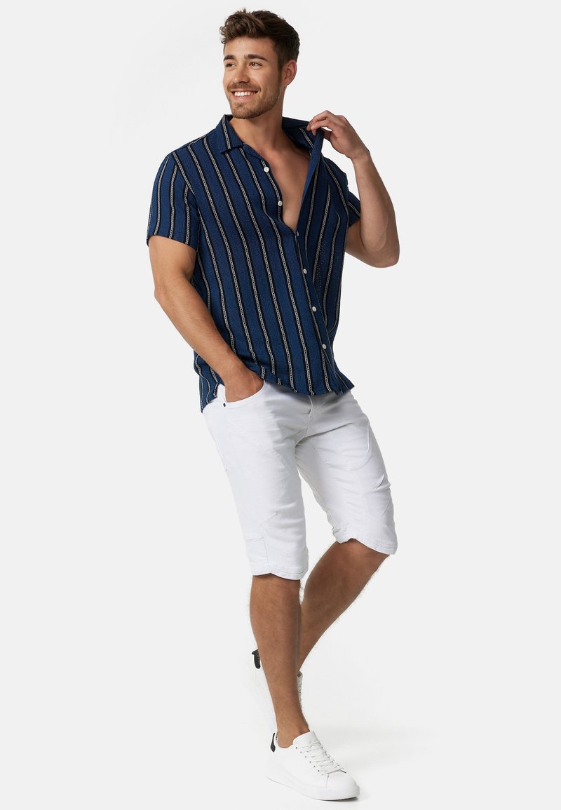 Chemise à manches courtes bleue avec des rayures verticales blanches et dorées, associée à un short blanc jusqu'au genou et des baskets blanches. Fond uni.