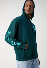 Felpa verde scuro in morbido tessuto con tasca frontale e cappuccio regolabile. Presenta una grafica floreale blu chiaro sulla manica destra e un'etichetta con logo.