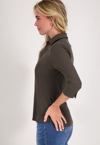 Donkerolijfgroene overhemd met kraag en driekwartmouwen, zijsplitten en een gladde stof; gedragen met blauwe jeans.