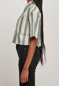 Persona con lunghi capelli intrecciati che indossa una camicia a maniche corte a righe verticali verde e bianca e pantaloni neri a vita alta, in piedi di profilo.
