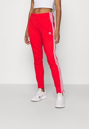 Kvinde iført røde Adidas træningsbukser med hvide sidelister og hvide sneakers, stående mod en ensfarvet lys baggrund.