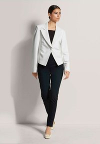 Weißer Blazer mit tailliertem Schnitt, einem einzigen Knopf, scharfen Revers und langen Ärmeln, kombiniert mit schwarzen Hosen. Das Model trägt beige Schuhe.
