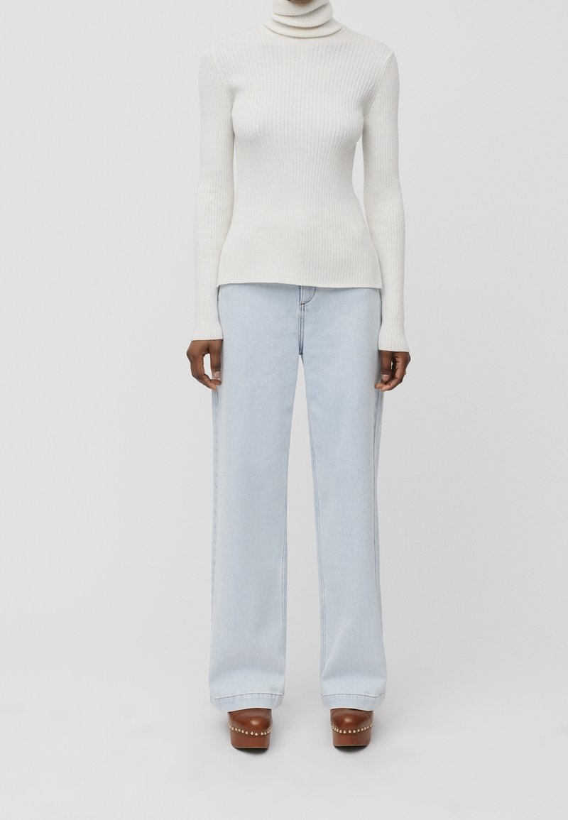 Col roulé blanc côtelé, silhouette ajustée. Jean bleu clair à jambes larges. Sabots marron ornés de clous argentés. Design simple et épuré avec un minimum de détails.