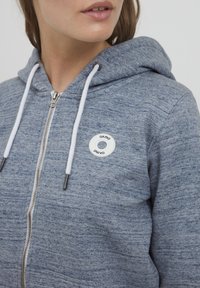 Grå zip-up hoodie tillverkad av texturerad tyg med vita dragsnören och en rund logopatch på bröstet. Dragkedjan sträcker sig till kragen.
