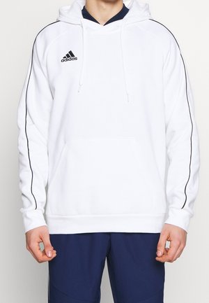 Personne portant un sweat à capuche blanc Adidas avec des bandes noires sur les manches et un pantalon bleu foncé, debout, les bras détendus.