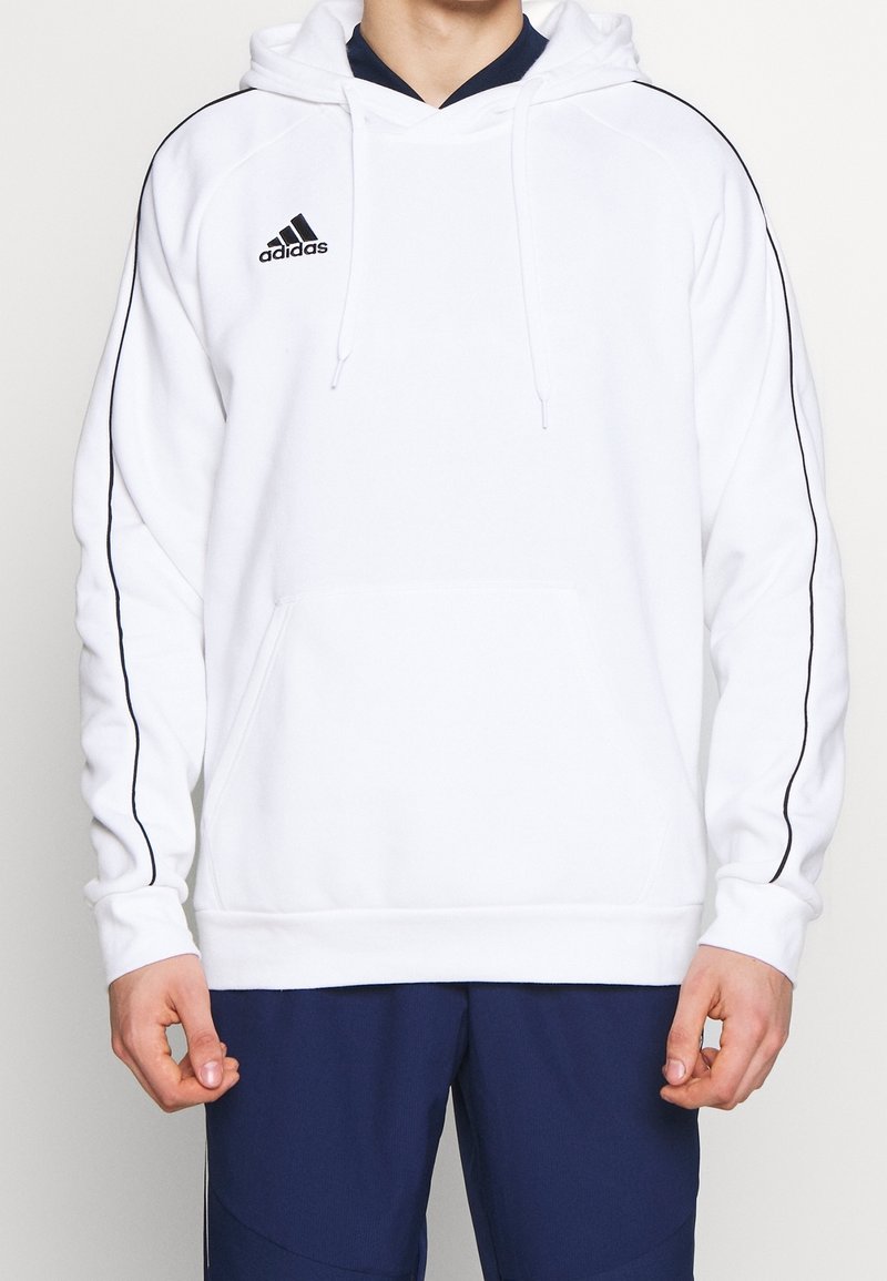 Personne portant un sweat à capuche blanc Adidas avec des bandes noires sur les manches et un pantalon bleu foncé, debout, les bras détendus.