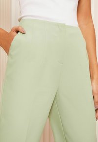 Pantaloni di un verde chiaro, su misura, realizzati in tessuto morbido, caratterizzati da pieghe frontali, tasche laterali e un design a gamba dritta.