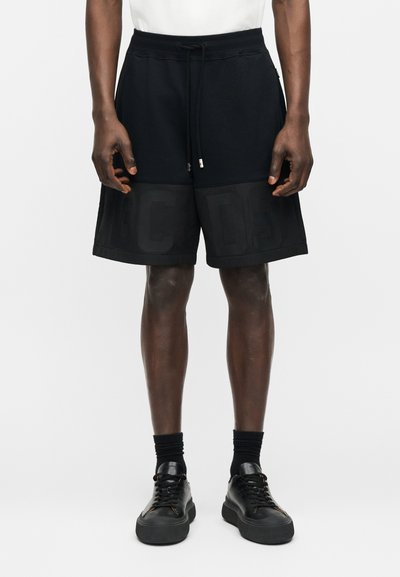 Άνδρας που φοράει μαύρα σορτς με κορδόνι και υφασμάτινα γράμματα "D S Q" κοντά στο τελείωμα, μαύρες κάλτσες και μαύρα αθλητικά παπούτσια σε λευκό φόντο.