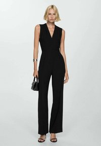 Svart ärmlös jumpsuit med djup v-ringning, skräddarsydd silhuett och vida ben; matchad med en liten svart handväska och bandade sandaler.
