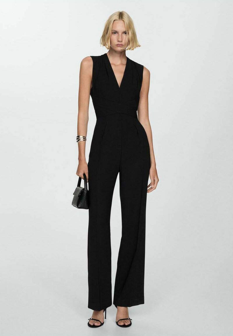 Svart ärmlös jumpsuit med djup v-ringning, skräddarsydd silhuett och vida ben; matchad med en liten svart handväska och bandade sandaler.