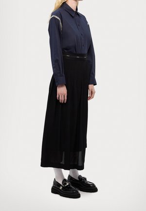 Femme portant une chemise bleu marine à manches longues boutonnée avec des détails en chaîne, une jupe midi noire, des chaussettes blanches et des mocassins noirs sur un fond uni.