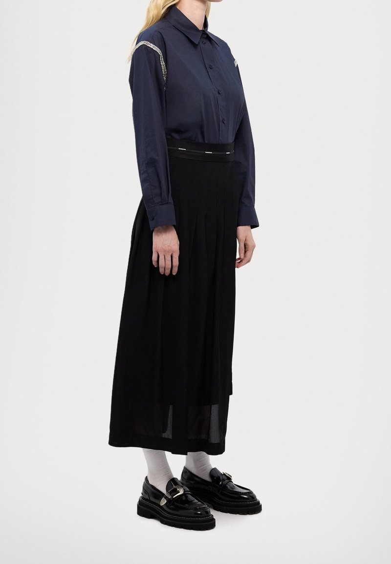 Femme portant une chemise bleu marine à manches longues boutonnée avec des détails en chaîne, une jupe midi noire, des chaussettes blanches et des mocassins noirs sur un fond uni.