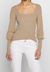 Beige, gerippter Strickpullover mit quadratischem Ausschnitt und langen, leicht puffigen Ärmeln; kombiniert mit weißen Jeans.