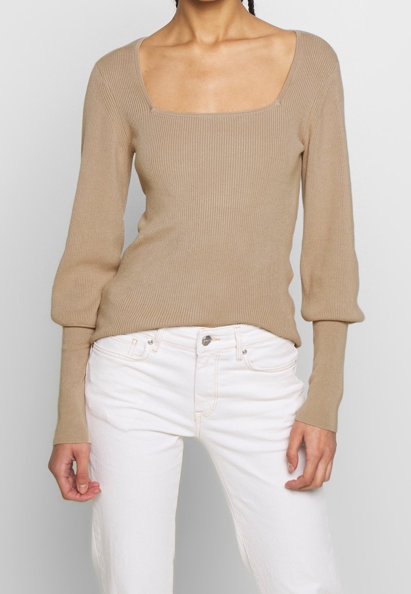 Beige, gerippter Strickpullover mit quadratischem Ausschnitt und langen, leicht puffigen Ärmeln; kombiniert mit weißen Jeans.