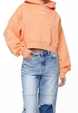 Personne portant un sweat à capuche court couleur pêche avec une poche avant et un jean en denim bleu bicolore délavé avec des détails déchirés.