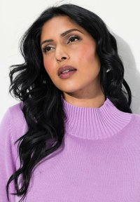 Hellblau gestrickter Pullover mit einem gerippten hohen Kragen. Das Model hat langes, gewelltes schwarzes Haar und ein natürliches Makeup, das ihre Gesichtszüge betont.