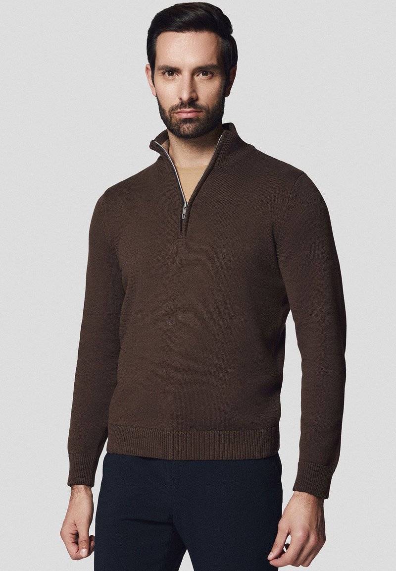 Brązowy sweter z kołnierzem na zamek, o dzianinowej teksturze, dopasowanym kroju i ściągaczach przy rękawach. Nosi się go na beżowej koszuli, w zestawieniu z ciemnymi spodniami.