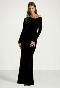 Kvinna som bär en lång svart sammetsklänning med off-shoulder som sitter tajt mot kroppen, stående mot en enkel ljus bakgrund.