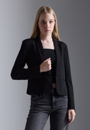 Black blazer made of smooth fabric with a fitted design, structured shoulders, and a single-button closure. Paired with a black top and jeans.