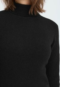 Donna che indossa un maglione lavorato a maglia nero con colletto alto, che mostra la texture e la vestibilità dalla nuca alla parte superiore del torso su uno sfondo neutro.