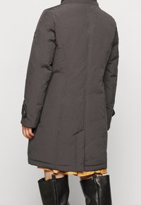 Person som bär en lång, mörk grå vadderad kappa med sömmar, en senapsgul kjol och svarta läderstövlar, sedd bakifrån.