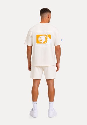 Uomo che indossa una maglietta bianca con grafiche arancioni di una tigre ruggente e testo, pantaloncini bianchi, calzini e scarpe da ginnastica, rivolto di spalle all'osservatore.