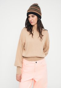 Μπεζ πουλόβερ με ψηλό λαιμό και φαρδιά μανίκια, σε συνδυασμό με ανοιχτό ροζ παντελόνι. Αξεσουάρ: πολύχρωμο πλεκτό καπέλο με ρίγες.