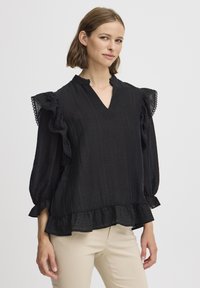 Blusa nera con texture, maniche a volant e scollatura a V. Presenta un bordo in pizzo e un orlo svasato. Indossata con pantaloni beige chiaro.