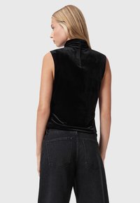 Blonde vrouw met de rug naar de camera, gekleed in een mouwloze zwarte fluwelen top met gerimpelde kraag en hoge zwarte denim jeans, tegen een grijze achtergrond.