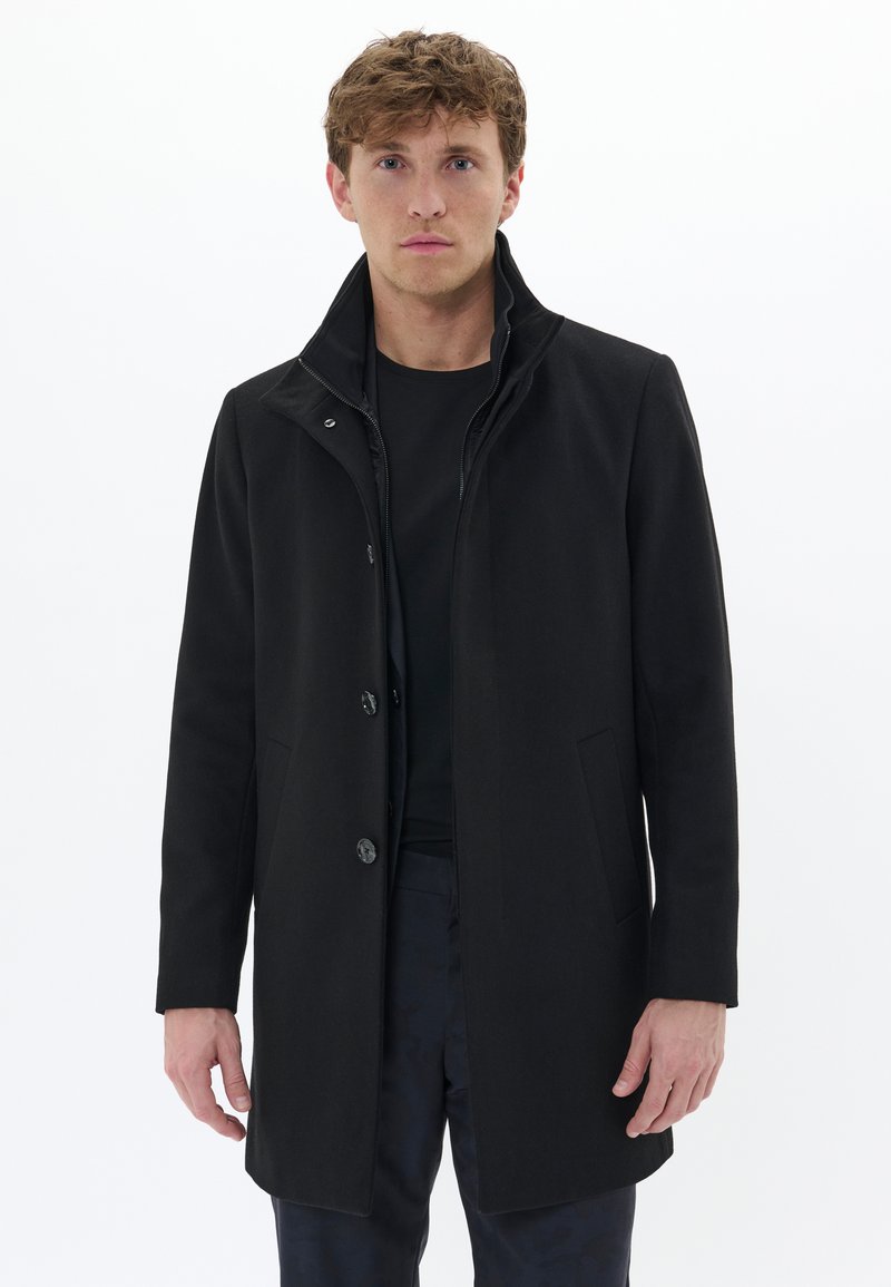 Cappotto nero con colletto alto, tessuto liscio, quattro bottoni, tasche laterali e una forma sagomata. Indossato sopra un top nero.