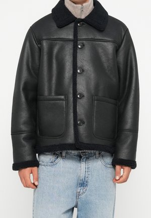 Czarna kurtka ze sztucznego baranka z dużymi guzikami, szerokim kołnierzem, przednimi kieszeniami, noszona na beżowym swetrze zapinanym na zamek i jasnoniebieskich dżinsach.