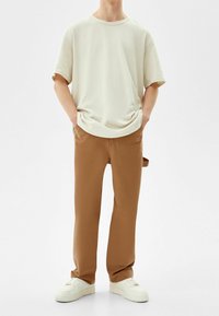 Jasno beżowa bawełniana koszulka o luźnym kroju i krótkich rękawach, w połączeniu z brązowymi spodniami straight-leg i białymi trampkami.
