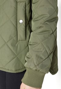 Quiltad grön jacka med en tryckknappsficka, ribbade ärmslut och ett strukturerat ytmaterial med ett diagonalt mönster.