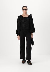 Schwarzes, strukturiertes Oberteil mit Puffärmeln, kombiniert mit schwarzen Jeans. Zubehör umfasst dunkle Sonnenbrillen und eine kleine schwarze Handtasche. Sandalen mit grobem Absatz.