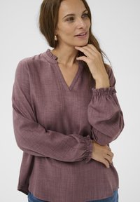 Femme aux longs cheveux bruns portant des boucles d'oreilles créoles en or et un chemisier mauve à manches longues avec un col en V et des poignets volantés, les bras croisés.