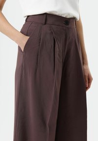 Wijd uitlopende broek in een diep bordeauxrode stof, met een gladde textuur, voorplooien en zijzakken. Afgewerkt met een knoopsluiting.