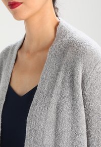 Grå texturerad kofta med öppen front, uppställd över en mörk v-ringad topp. Mjuk väv med en mysig känsla, som har en lätt design.