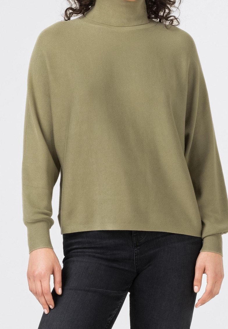 Woman wearing an olive green turtleneck sweater and black jeans, standing with arms relaxed by her sides.