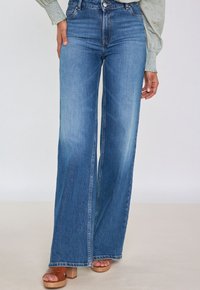 Jeans in denim blu a vita alta e svasati con una finitura scolorita. Presentano una chiusura a bottone e due tasche frontali. Abbinati a sandali con plateau marroni.