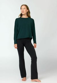 Frau steht barfuß und trägt ein dunkelgrünes Langarmshirt sowie eng anliegende schwarze Yogahosen vor einem schlichten hellen Hintergrund.