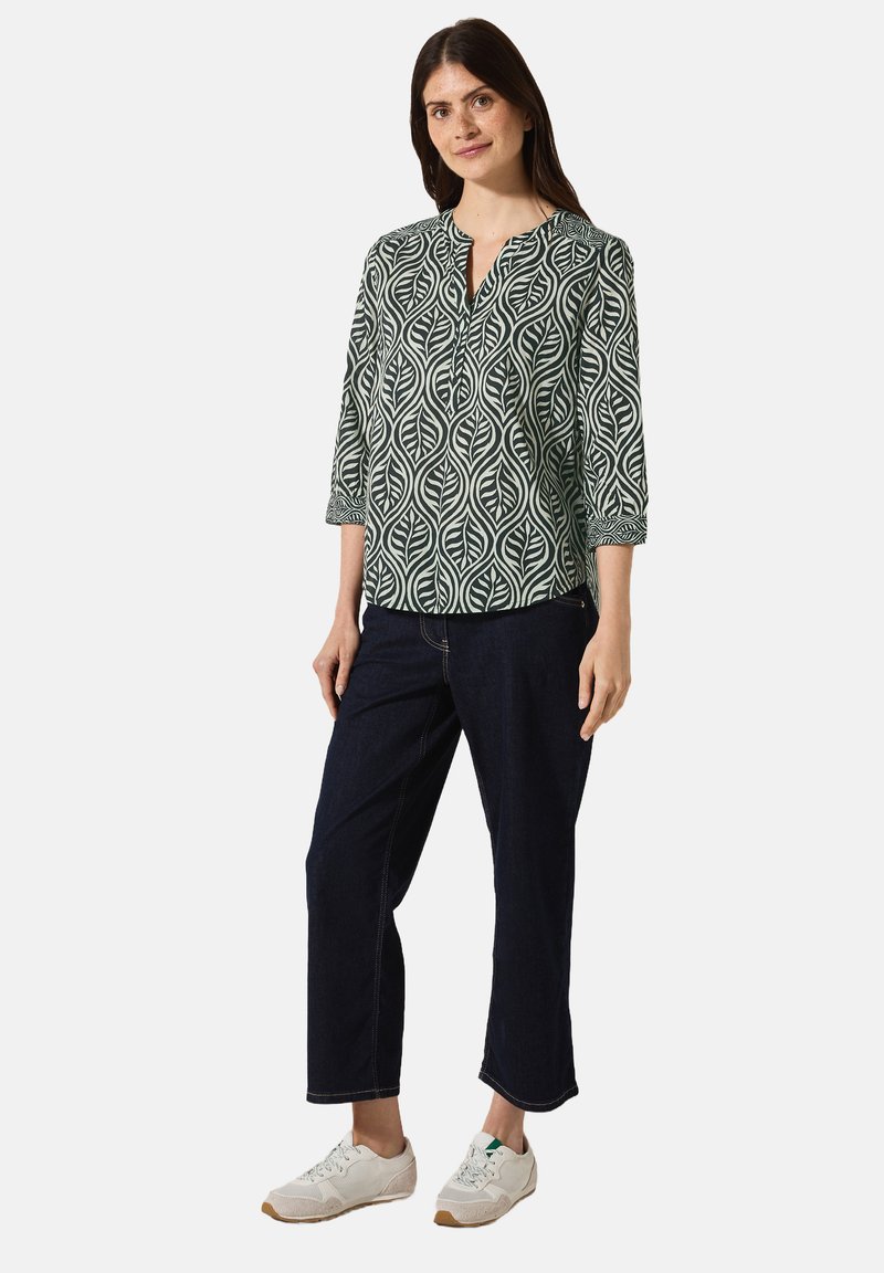 Groen en crèmekleurig blouse met een bladdessin, v-hals en driekwart mouwen, gecombineerd met donkere jeans en witte sneakers.
