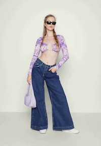 Top de mangas longas com padrão de mármore roxo e recorte em forma de olho-chave, combinado com jeans de denim azul de perna larga e uma mala de ombro lavanda.