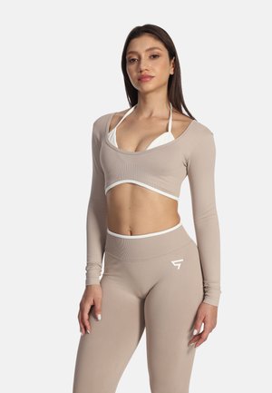 Žena oblečená v béžovom dlhom crop tope cez biely bikiny top a legíny s vysokým pásom stojí pred svetlým jednofarebným pozadím.