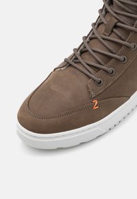 Bruine leren hoge sneakers met grijze veters, voorzien van oranje accenten en een witte rubberen zool. Glad oppervlak en gedetailleerde stiksels.