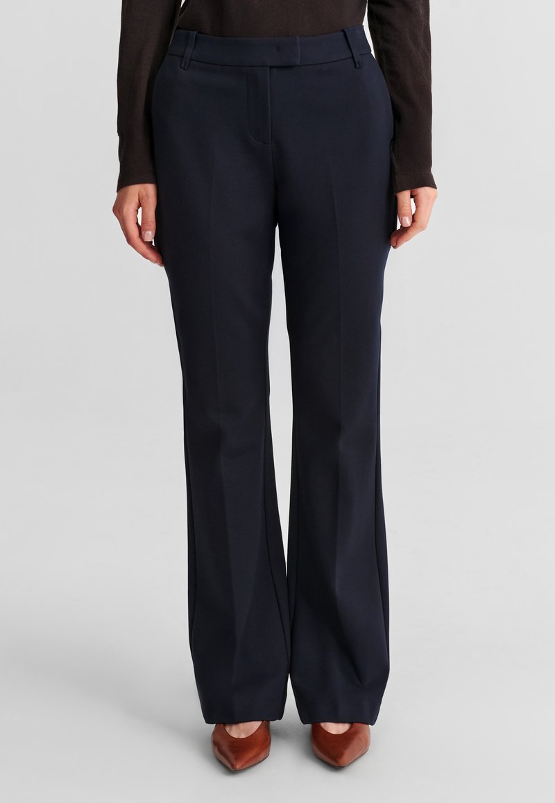 Femme portant un pantalon large bleu marine sur mesure et des talons pointus marron, debout devant un fond gris.