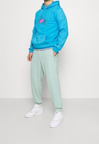 Man som bär en ljusblå hoodie med rosa grafik, ljusgröna sweatpants och vita Adidas-sneakers, står med en hand i fickan.